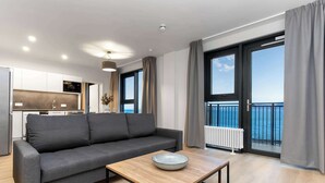 Una sala de estar moderna con un sofá gris, una mesa de centro de madera y vistas al mar a través de ventanas grandes.