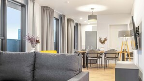 Ein modernes Wohnzimmer mit grauem Sofa, Essbereich mit Stühlen und Tisch sowie einem an der Wand befestigten Fernseher.