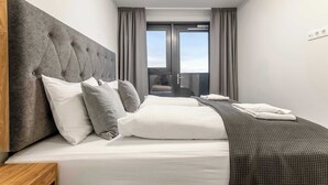Ein modernes Hotelzimmer mit einem großen Bett, einer grauen Kopfteil und Blick durch eine Glastür.