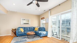 A living room with a blue sectional sofa, a round coffee table, and a large window with a view of a deck. - Cute, spacious 4-bedroom townhouse with free WiFi and easy access to shops located downtown on the ocean block! (Ocean City)
