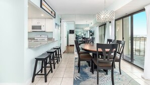 Un espace repas moderne avec une grande table et des chaises, une cuisine dotée de meubles blancs et une vue sur l’extérieur grâce à une porte vitrée.
