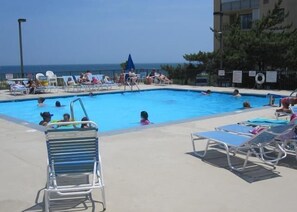 A swimming pool with lounge chairs and umbrellas, surrounded by a fence and trees. - Cozy 2 Bedroom Oceanfront Condo with Outdoor Pool, Coastal Decor, and Amazing View is Perfect for Families! (Ocean City)