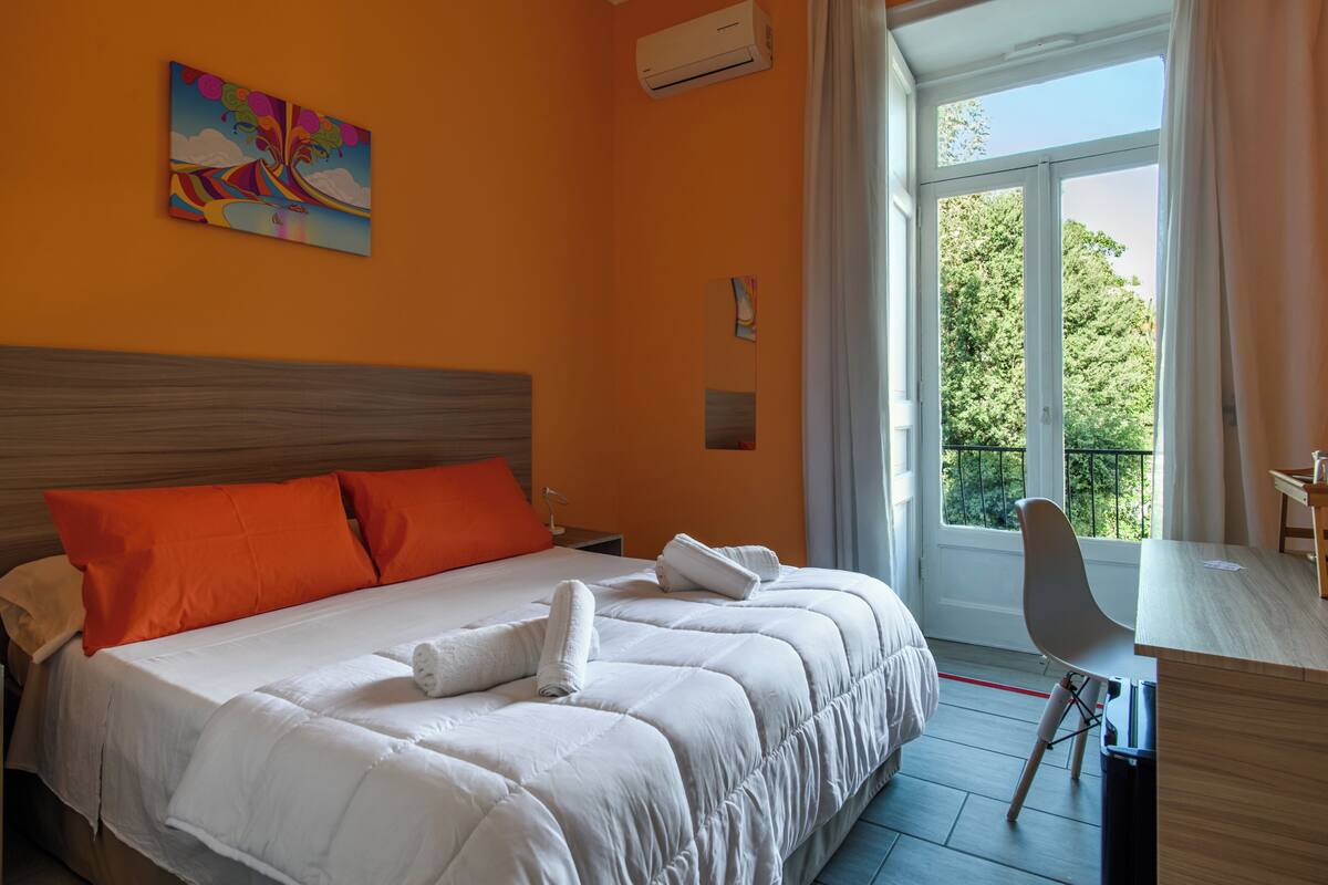 A hotel room with a bed, orange pillows, a desk, and a window with a view of trees.