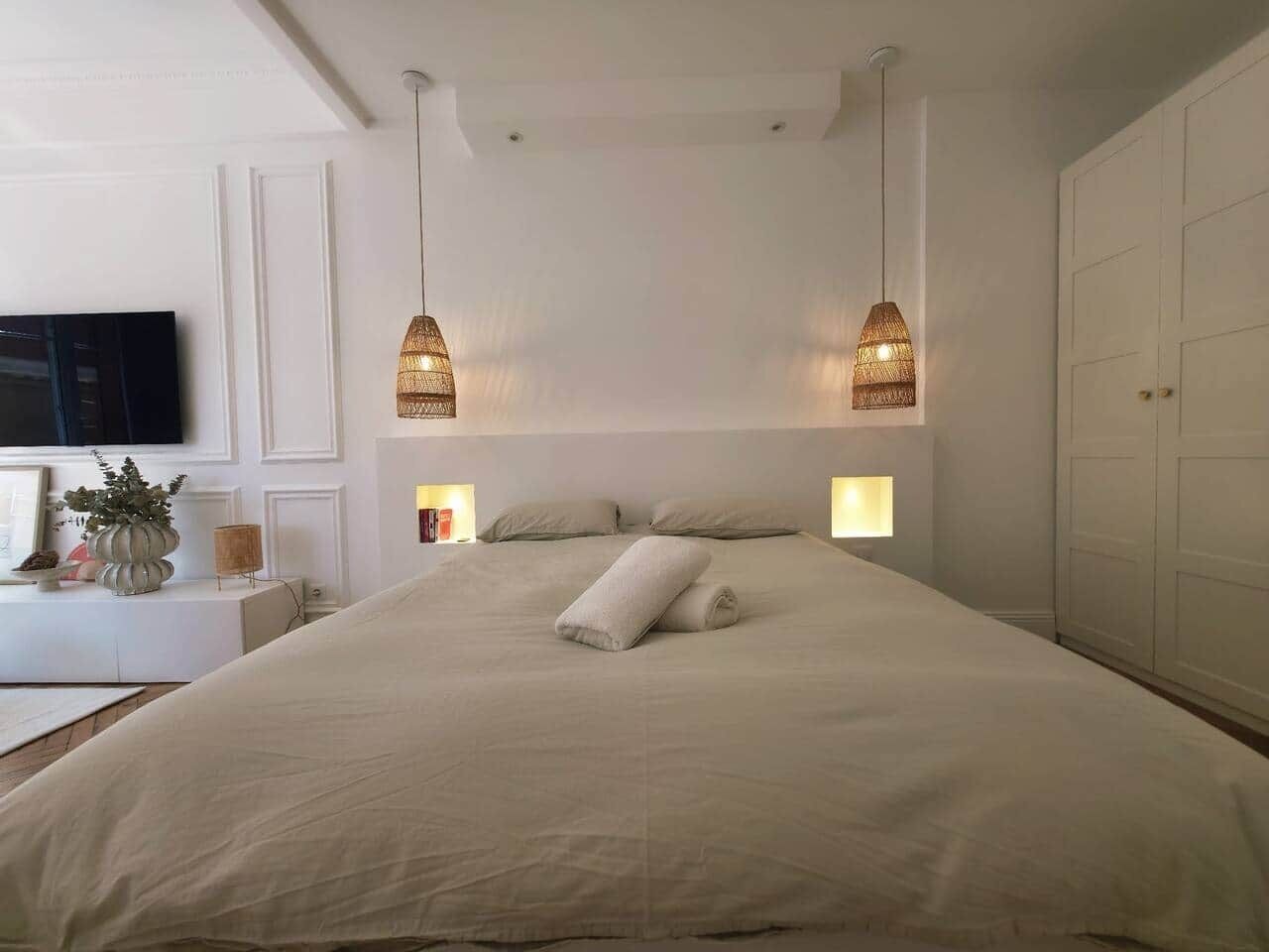 A neatly made bed with two pillows, a bedside table with a lamp, and a television on the wall.