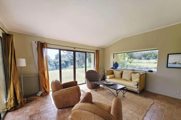 A living room with a sofa, armchairs, a coffee table, and a view of a garden through large windows. - Magnificent villa for 14 people, with a pool, garden, and parking (La Colle-sur-Loup)