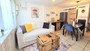 A living room with a sofa, a wooden chest, a dining table with chairs, a kitchen area, and a hanging light fixture. - Loù Cabanoù 35 m from the seafront (Marseille)