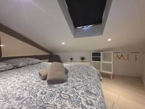 Ein Schlafzimmer mit Dachfenster, einem Bett mit gemusterten Betttextilien, einem Nachttisch, einer kleinen Pflanze und einem Kleiderschrank.