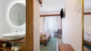 Une chambre d’hôtel avec un miroir rond, un lavabo, un lit recouvert d’une courtepointe à motifs floraux, une chaise et un téléviseur fixé au mur.