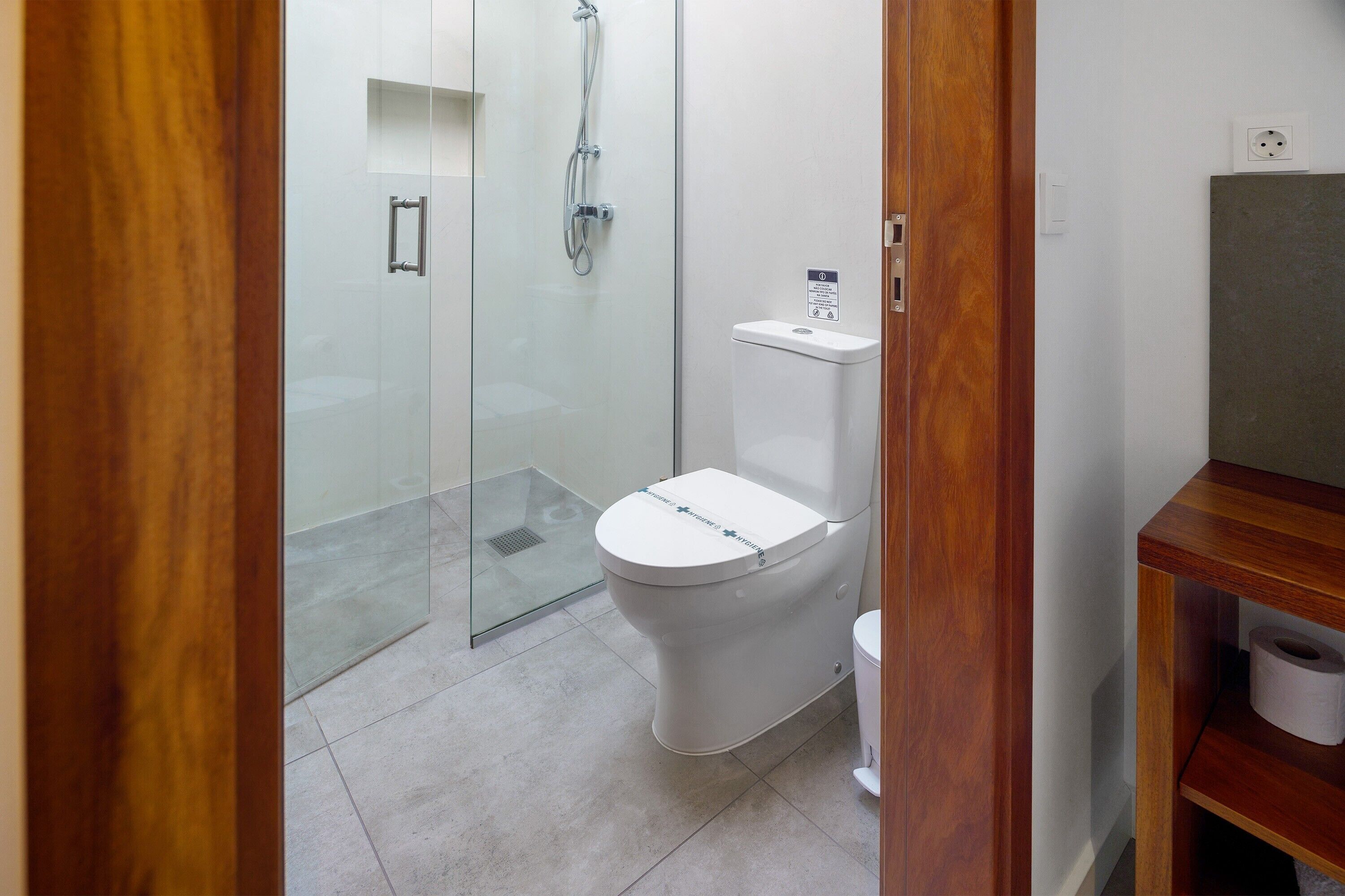 Een badkamer met een glazen douchecabine, een toilet en een houten wastafelmeubel met een plank.