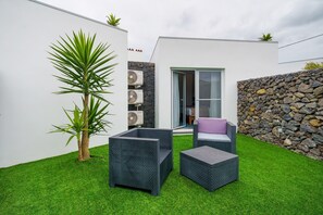 Een modern terras buiten, met een stenen muur, een palmboom en twee rieten stoelen.