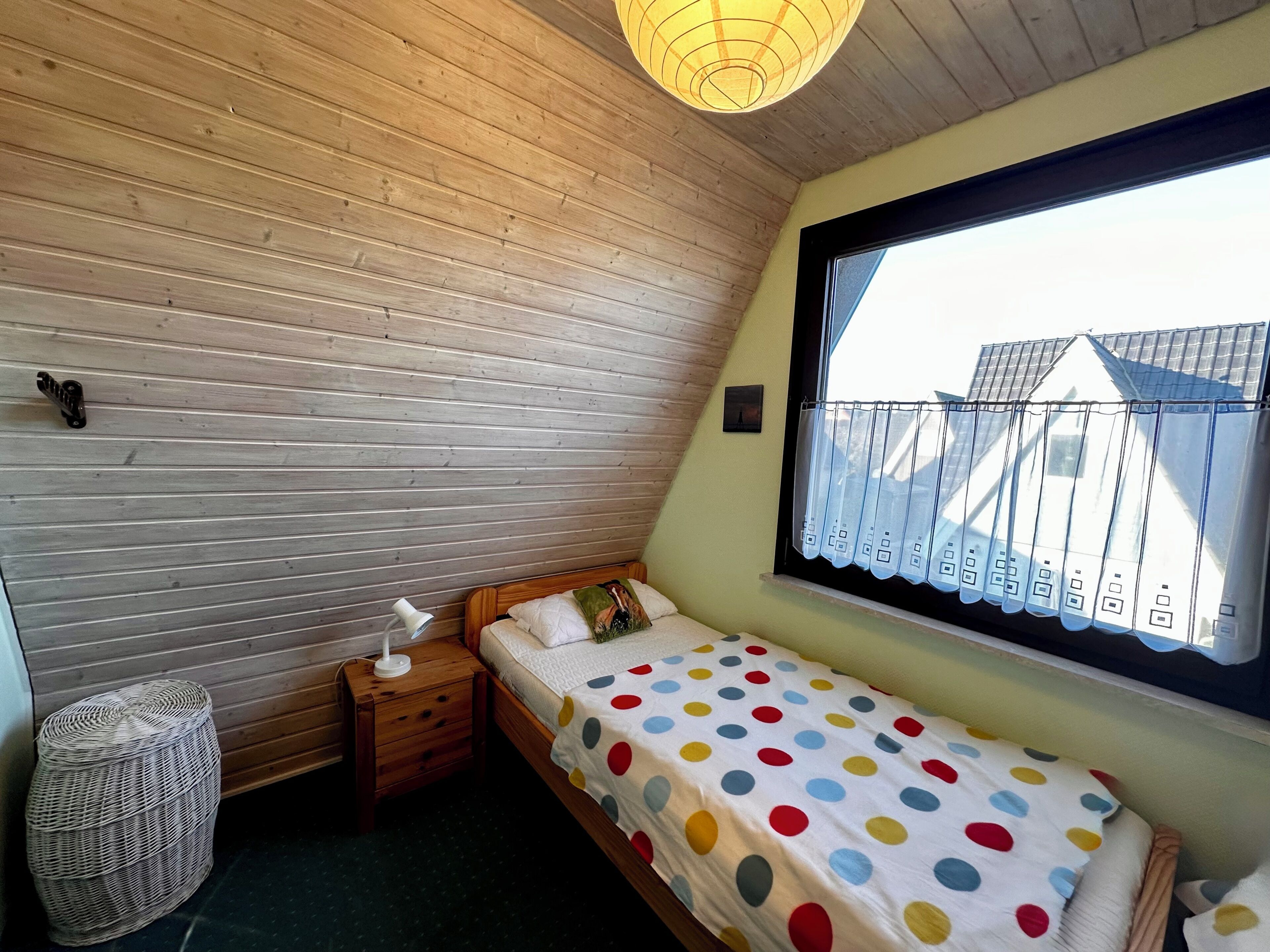 Un dormitorio pequeño con techo de listones de madera, una cama individual con colcha de lunares, una mesita de noche con lámpara y una ventana con cortina.