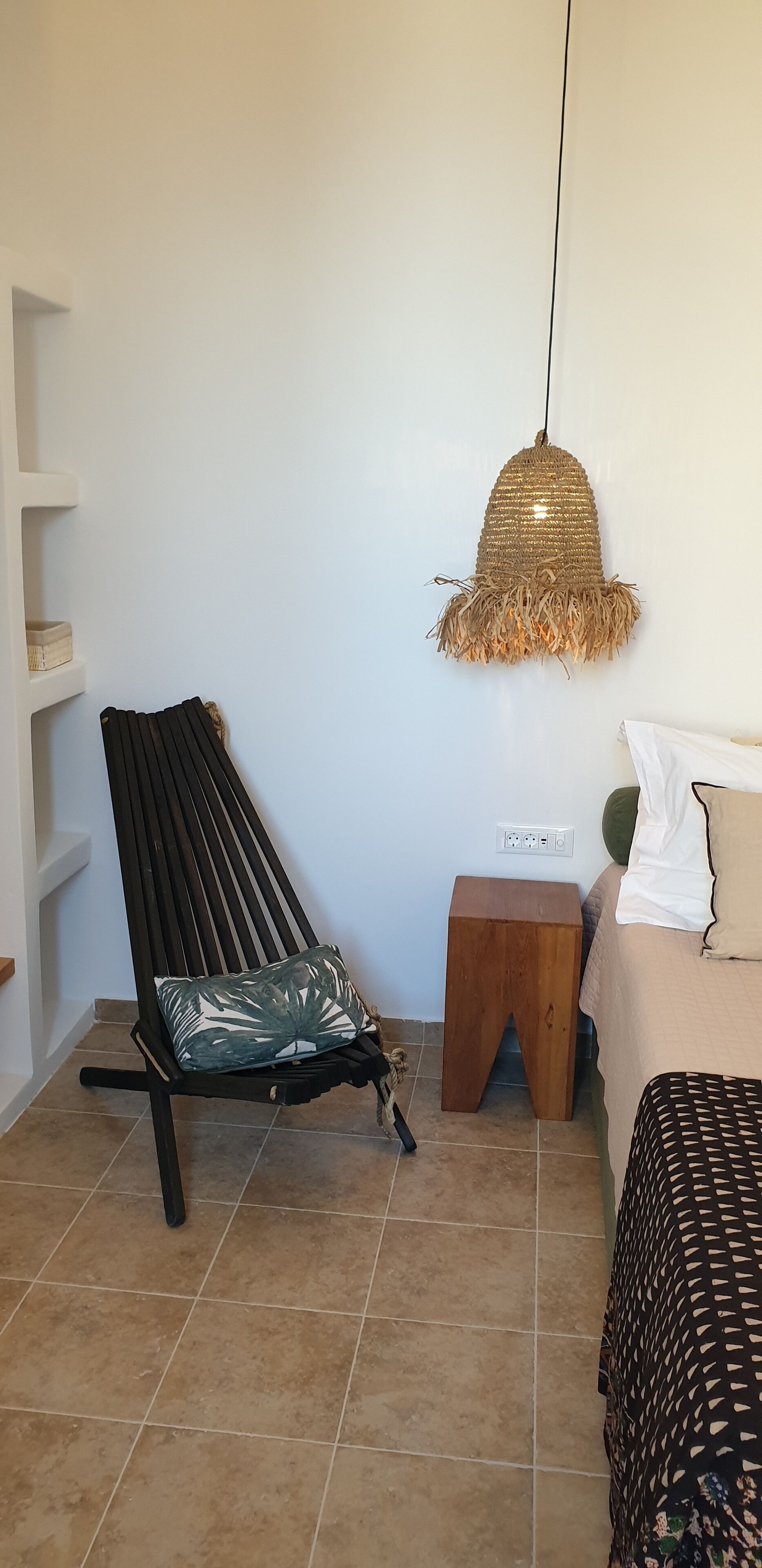 A room with a black lounge chair, a small wooden stool, a woven pendant light, and a bed with patterned bedding.