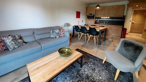 Un salon moderne avec un canapé gris, une table basse en bois et un fauteuil gris. On y trouve également un coin repas avec une table et des chaises en bois, et une cuisine à l’arrière-plan.