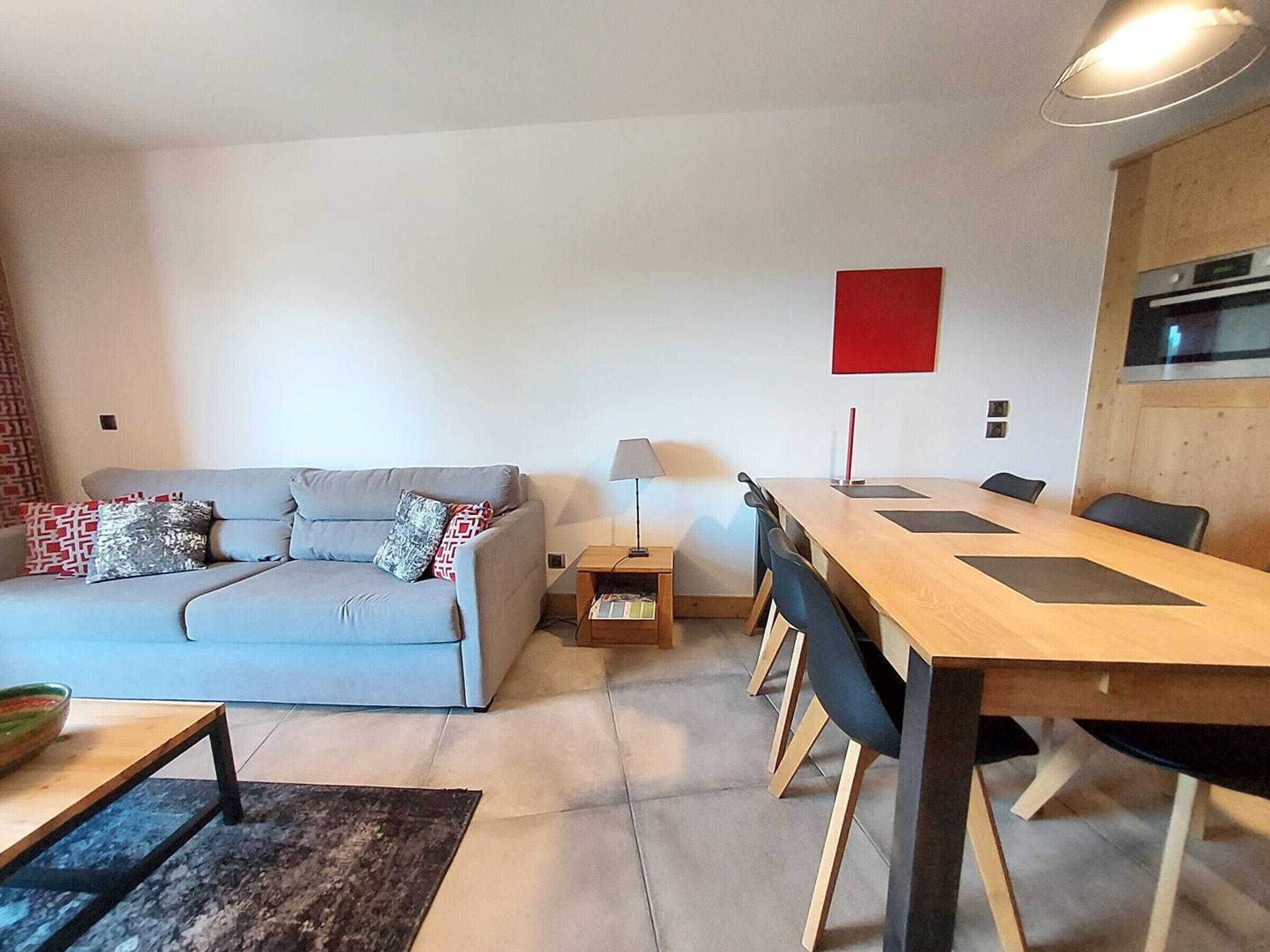 A modern living room with a grey sofa, a wooden dining table, and a red accent wall.