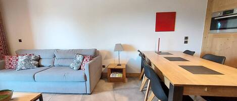 A modern living room with a grey sofa, a wooden dining table, and a red accent wall.
