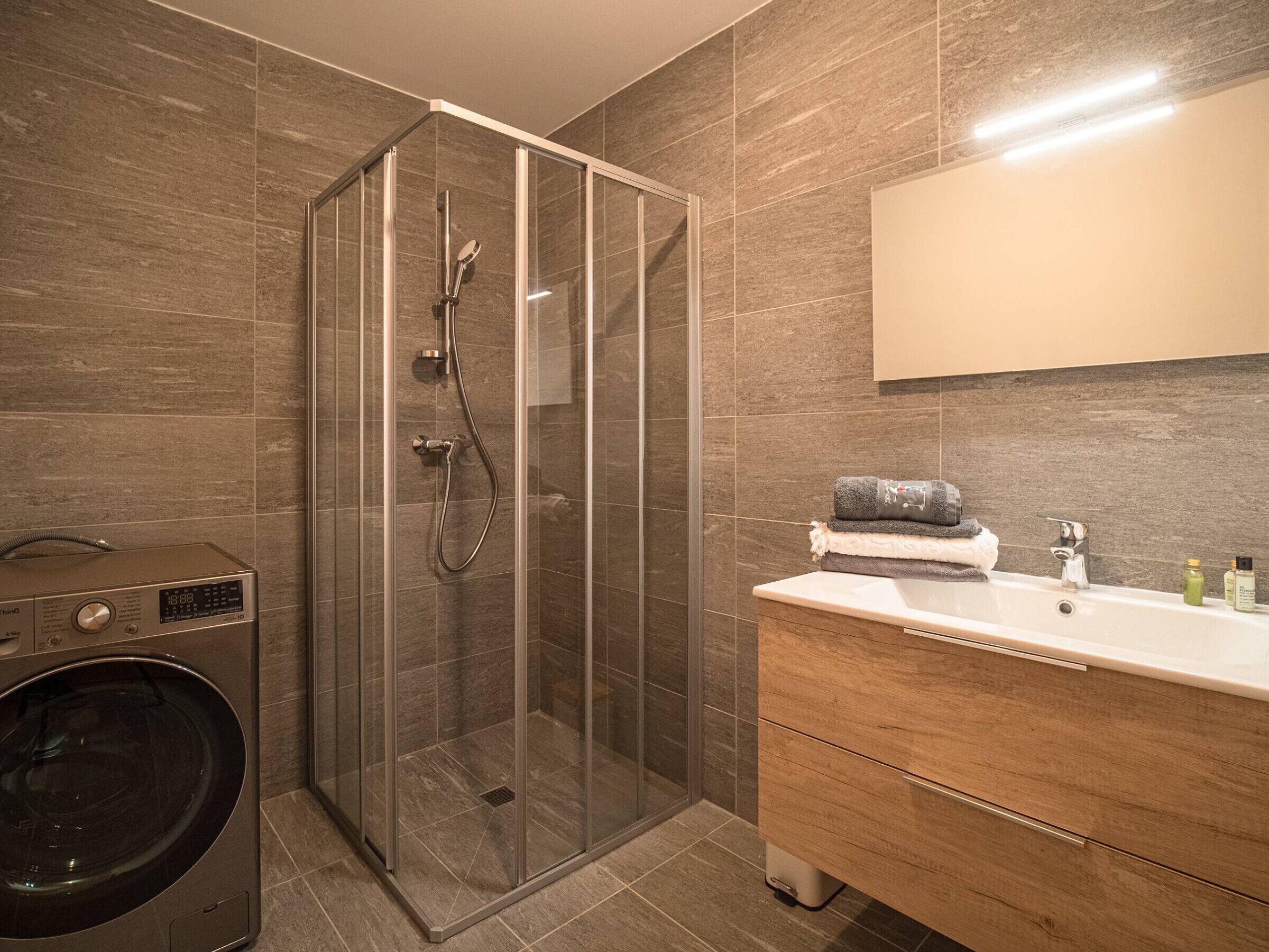 A modern bathroom with a glass-enclosed shower, a built-in washing machine, and a vanity with a mirror and toiletries.
