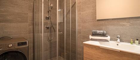 A modern bathroom with a glass-enclosed shower, a built-in washing machine, and a vanity with a mirror and toiletries.