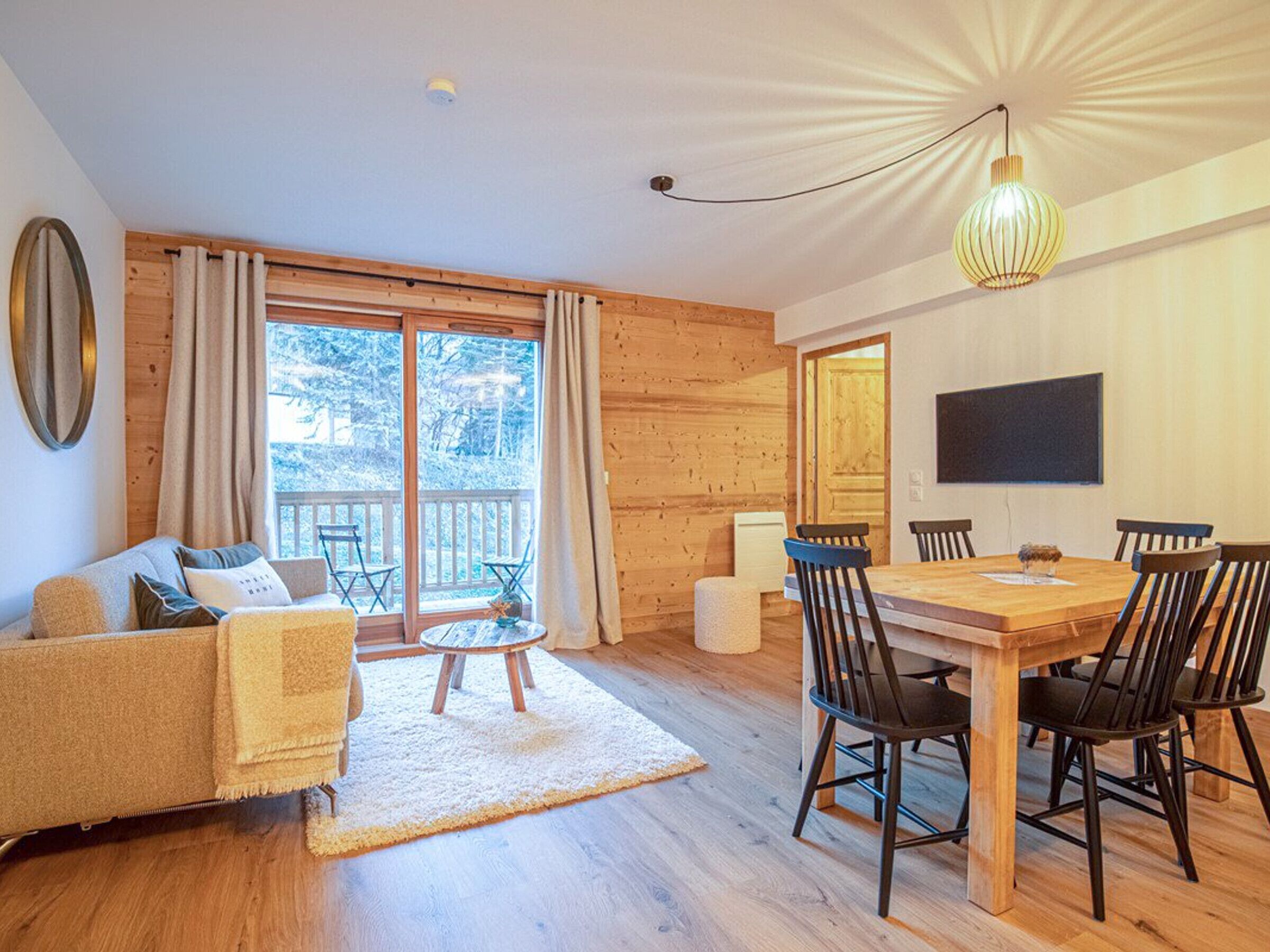 A cozy room with a wooden floor, a sofa, a dining table with chairs, and a large window with curtains.