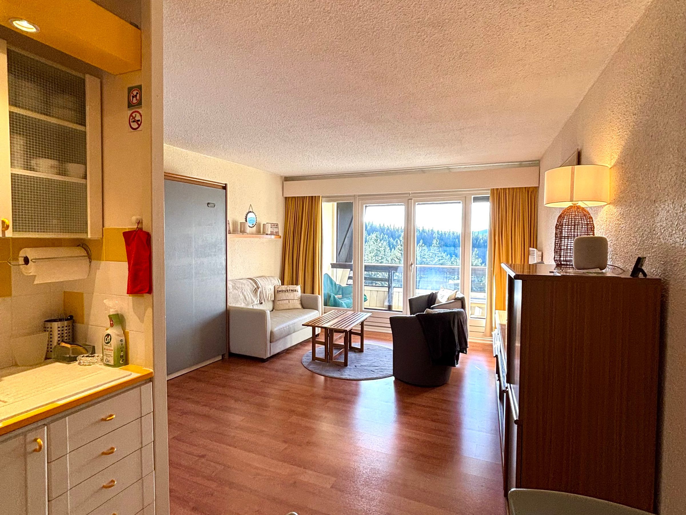 Een ruime woonkamer met een bank, een bijzettafeltje en een balkon met uitzicht op bomen.