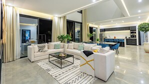 A modern living room with a sectional sofa, a glass coffee table, and a dining area with blue chairs. - Beautiful villa with swimming pool at Argan Golf Resort (Marrakesh)