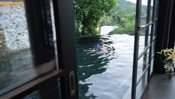 Vista de uma piscina com uma criança nadando, cercada por vegetação e um muro de pedra.