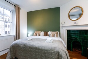 Une chambre à coucher avec un lit, des oreillers, une lampe de chevet, un miroir et une cheminée recouverte de carreaux verts.