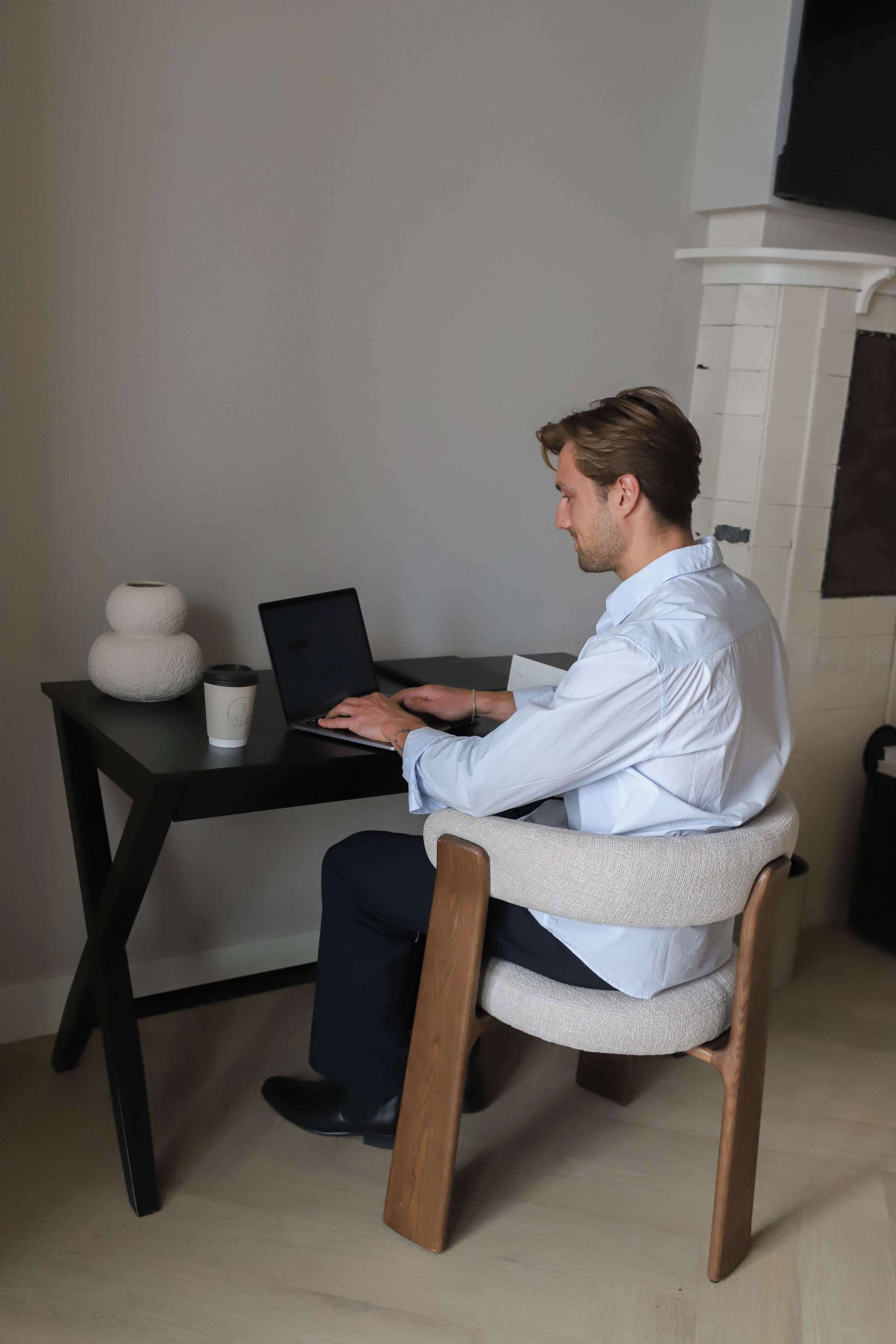 Iemand zit aan een zwarte tafel met een laptop, een witte mok en een witte vaas.