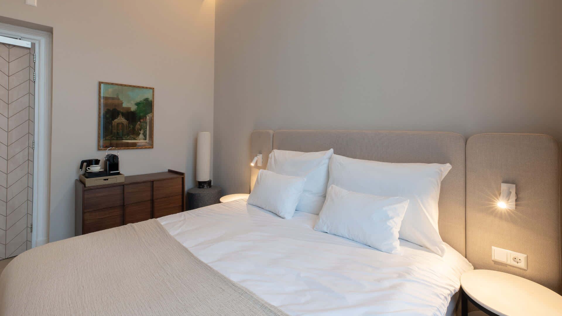 Un letto rifatto con lenzuola bianche, un comodino con una lampada e un quadro appeso al muro.