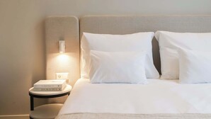 Ein ordentlich gemachtes Bett mit weißen Leinen und Kissen, ein Nachttisch mit einem Buch und einer Lampe sowie eine Wandleuchte.