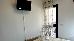 Et hotelværelse med en seng, et fjernsyn ophængt på væggen, et vindue med gardiner og et håndklædetørrestativ.