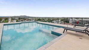 Une piscine sur le toit, avec une eau d’un bleu limpide, entourée d’une rambarde en métal et de quelques chaises longues.