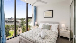 Une chambre d’hôtel avec un grand lit, un ventilateur de plafond, une vue sur la ville et un tableau encadré au mur.