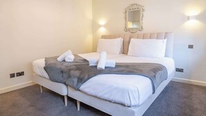 Un lit bien fait, avec du linge de lit blanc et un plaid gris, un miroir fixé au mur et deux appliques murales qui diffusent la lumière.