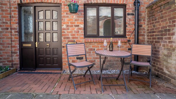 Un bâtiment en briques avec une porte marron, deux chaises en métal et une table sur laquelle se trouve une bouteille.