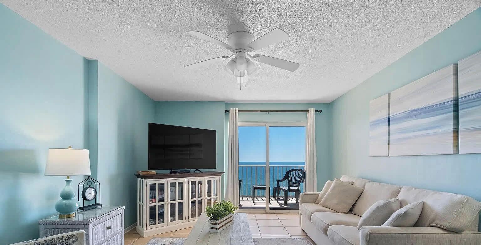 A living room with a ceiling fan, a flat-screen TV, a sofa, a coffee table, and a view of the ocean through a sliding glass door.