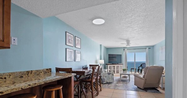 A spacious living area with a dining table, chairs, a TV, and a balcony with a view of the ocean. - 2B 2B Beach Views Best Eats Palmaway (Gulf Shores)