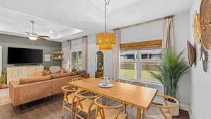 Ein modernes Wohnzimmer mit einer Couch, einem Esstisch und Stühlen. Es gibt einen Fernseher, einen Deckenventilator und ein großes Fenster mit Blick auf den Garten.