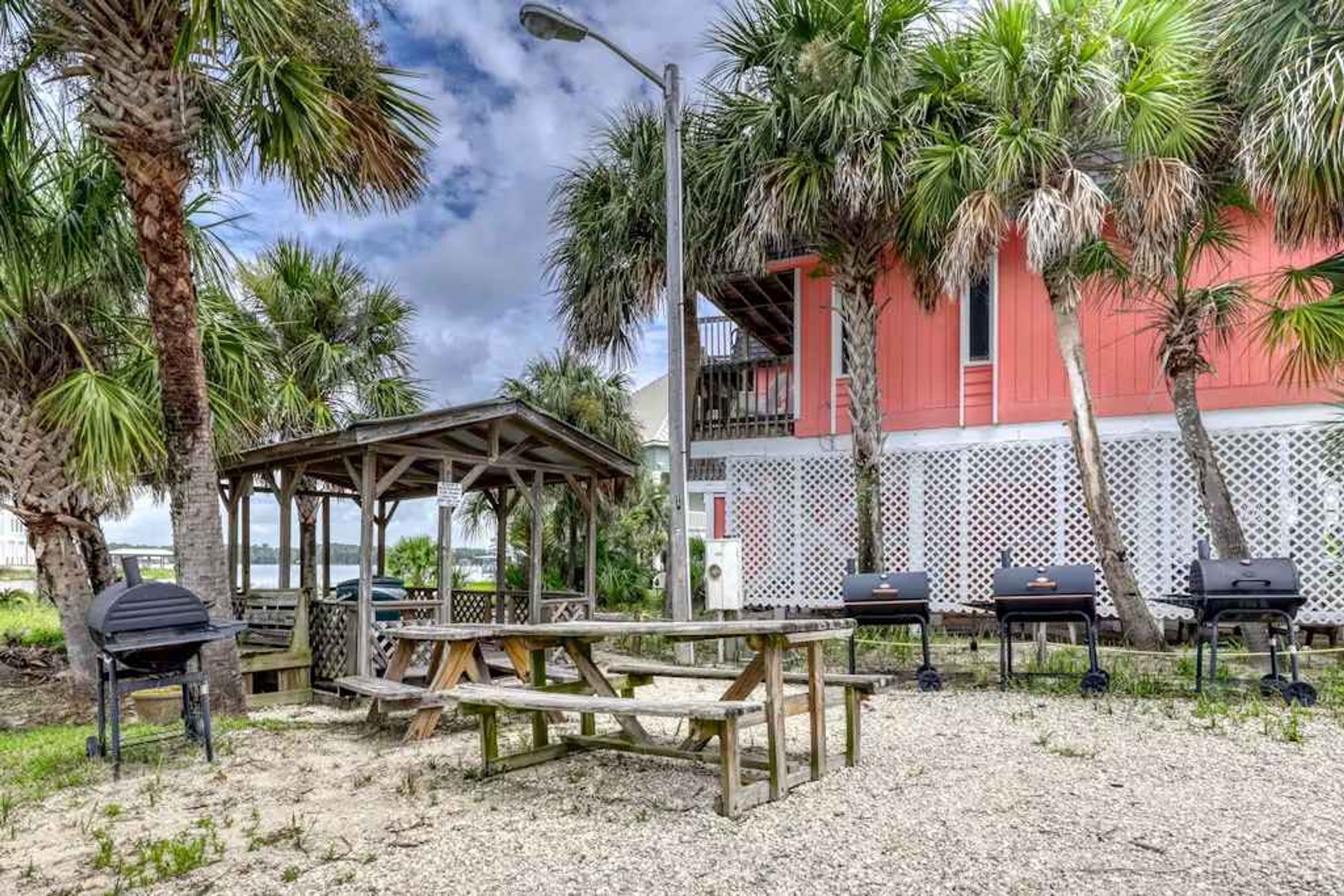 A beachside patio with a wooden gazebo, barbecue grills, and a red building with a balcony.