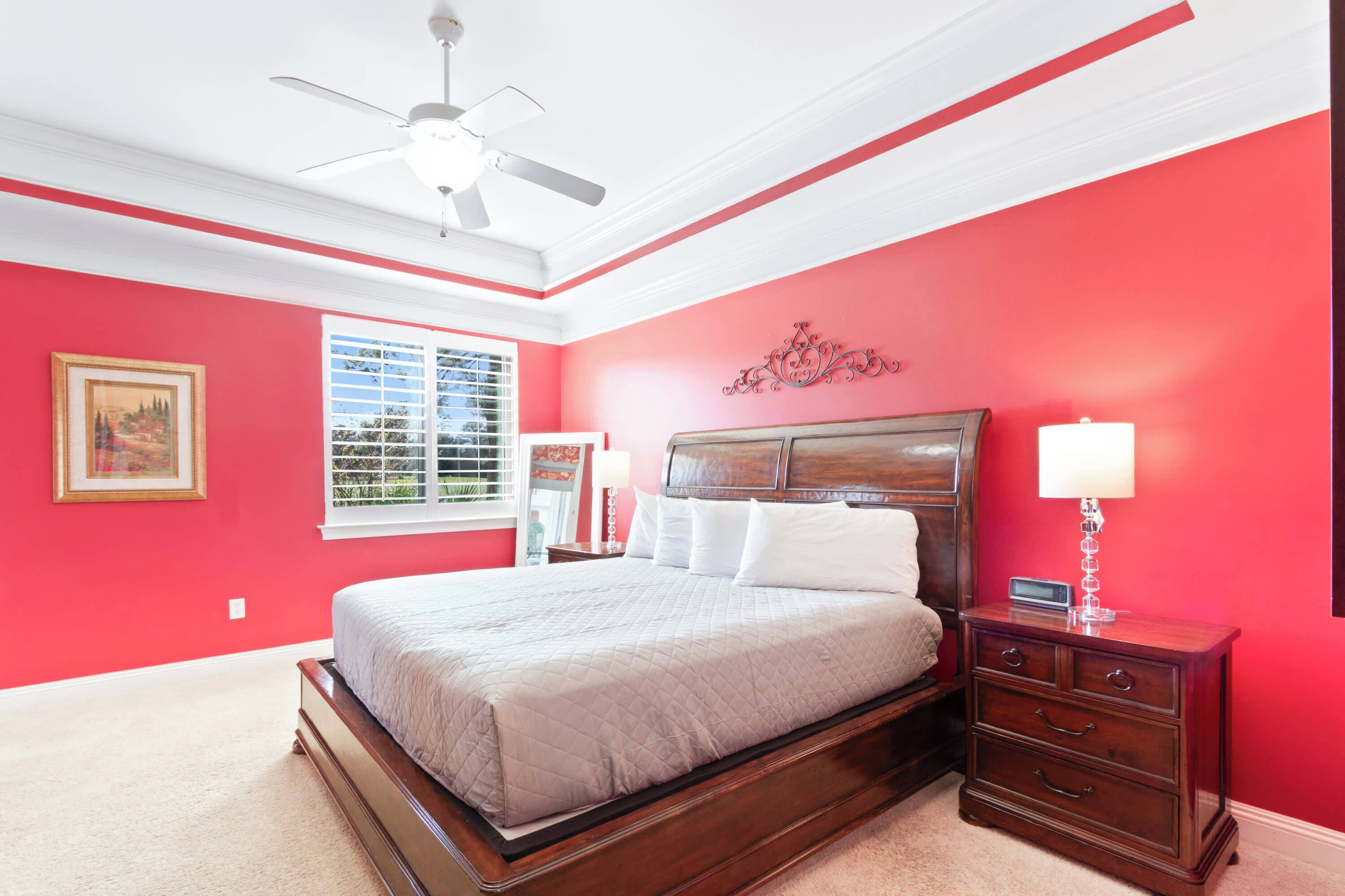 Une chambre à coucher comprenant un lit, une table de chevet, une commode, un ventilateur de plafond et une fenêtre avec des stores.