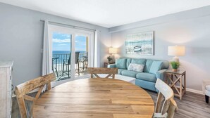 A living room with a wooden dining table, a blue sofa, and a view of the ocean through a sliding glass door. - NEW Gulf Front - Beaches Pool Dining and Fun (Gulf Shores)