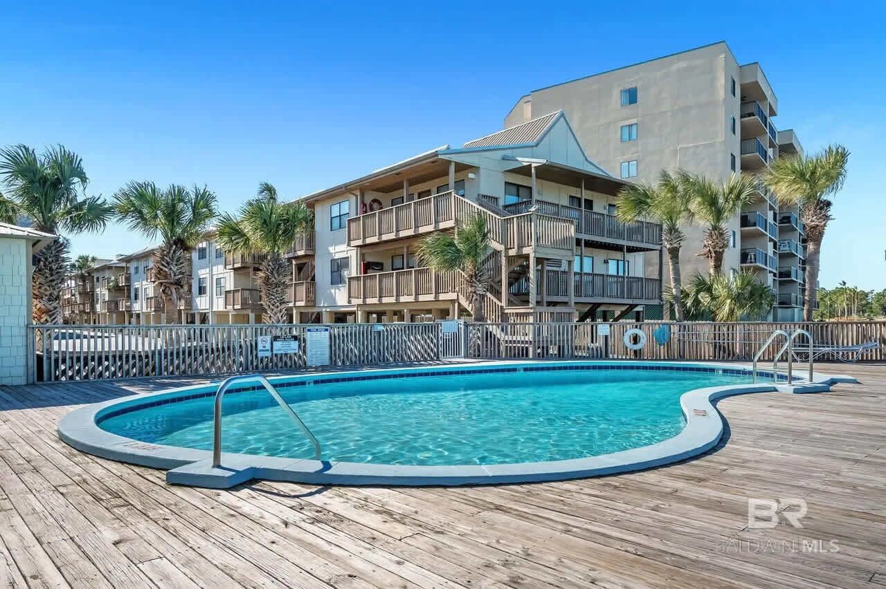 A pool area with a wooden deck, surrounded by multi-story buildings with balconies and palm trees.