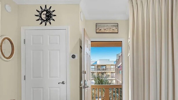 A hallway with a white door, a wooden table with a bowl, a sun decoration, and a view of a building through an open door. - 1 bed Beach Pools Waterfront Boating Palmaway (Orange Beach)