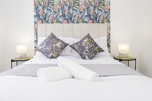 A neatly made bed with white linens, two decorative pillows with a marble pattern, and a floral tapestry headboard. There are two bedside tables with lamps and candles. - Stunning Home in Balsall Heath With Patio and Central Heating (Birmingham)