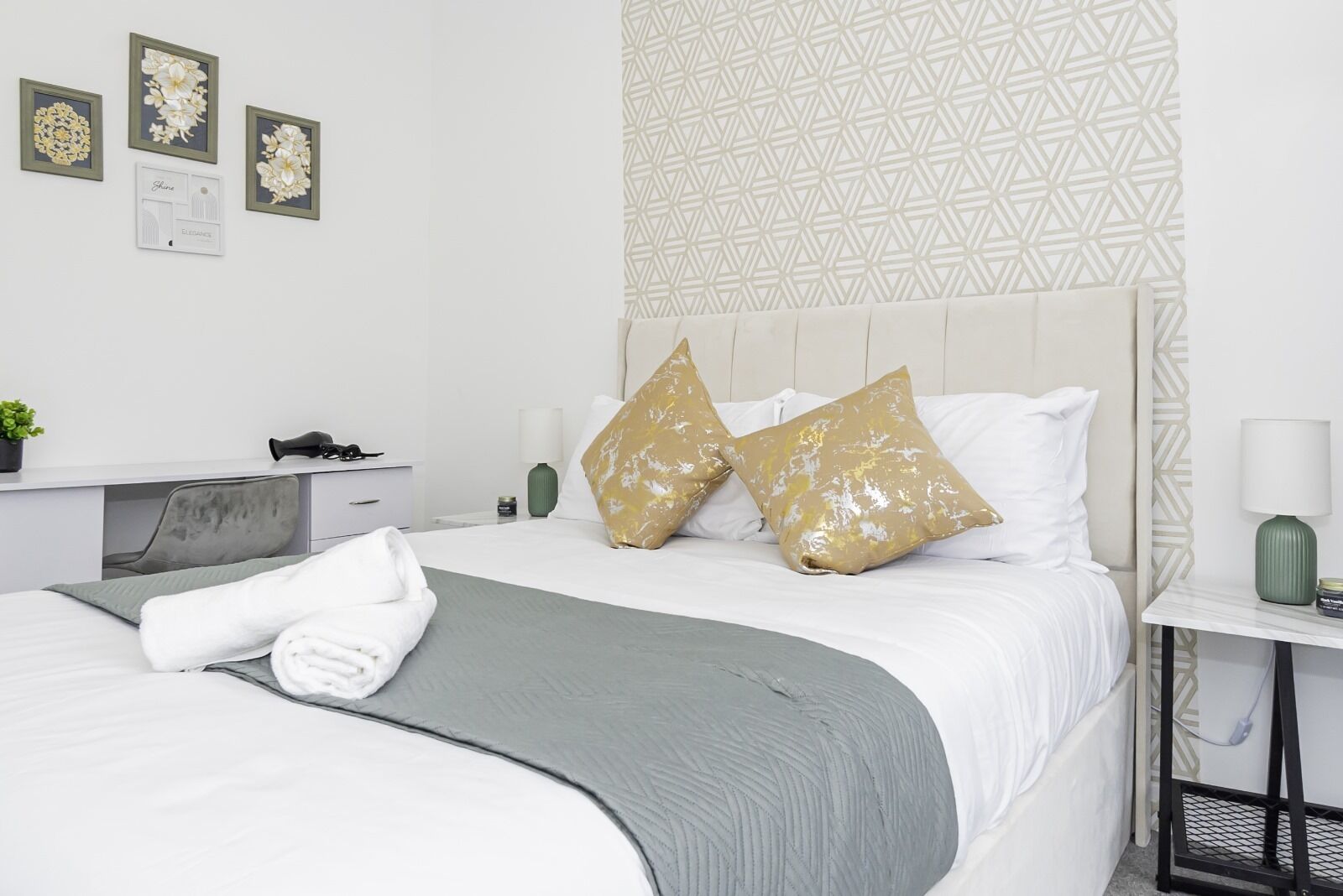 A neatly made bed with a grey blanket, two gold pillows, and a white pillow. There is a small bedside table with a green vase and a lamp. The wall features a geometric pattern and three framed floral artworks.