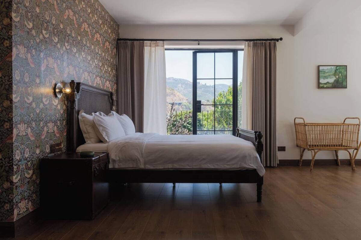 Un dormitorio con una cama grande, una mesita de noche, una silla de mimbre y vistas al exterior a través de una ventana.