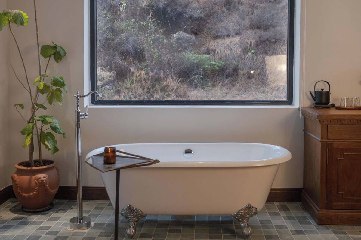 Un baño con una bañera independiente, una ventana con vistas a un paisaje natural, una planta en maceta y un armario de madera.