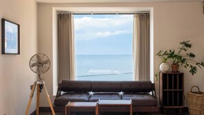 Ένα σαλόνι με ένα μεγάλο παράθυρο, έναν καφέ δερμάτινο καναπέ, ένα ξύλινο τραπεζάκι σαλονιού, έναν ανεμιστήρα δαπέδου, ένα φυτό σε γλάστρα και ένα καλάθι από ρατάν.