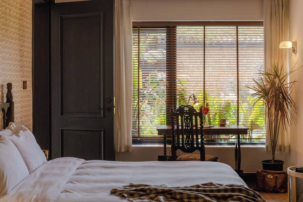 Un dormitorio con cama, ventana con persianas, una mesa con silla y una planta en maceta.