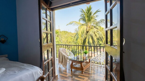 A bedroom with a wooden bed, a balcony with a view of palm trees, and a blue wall. - Celeste Stays (Candolim)