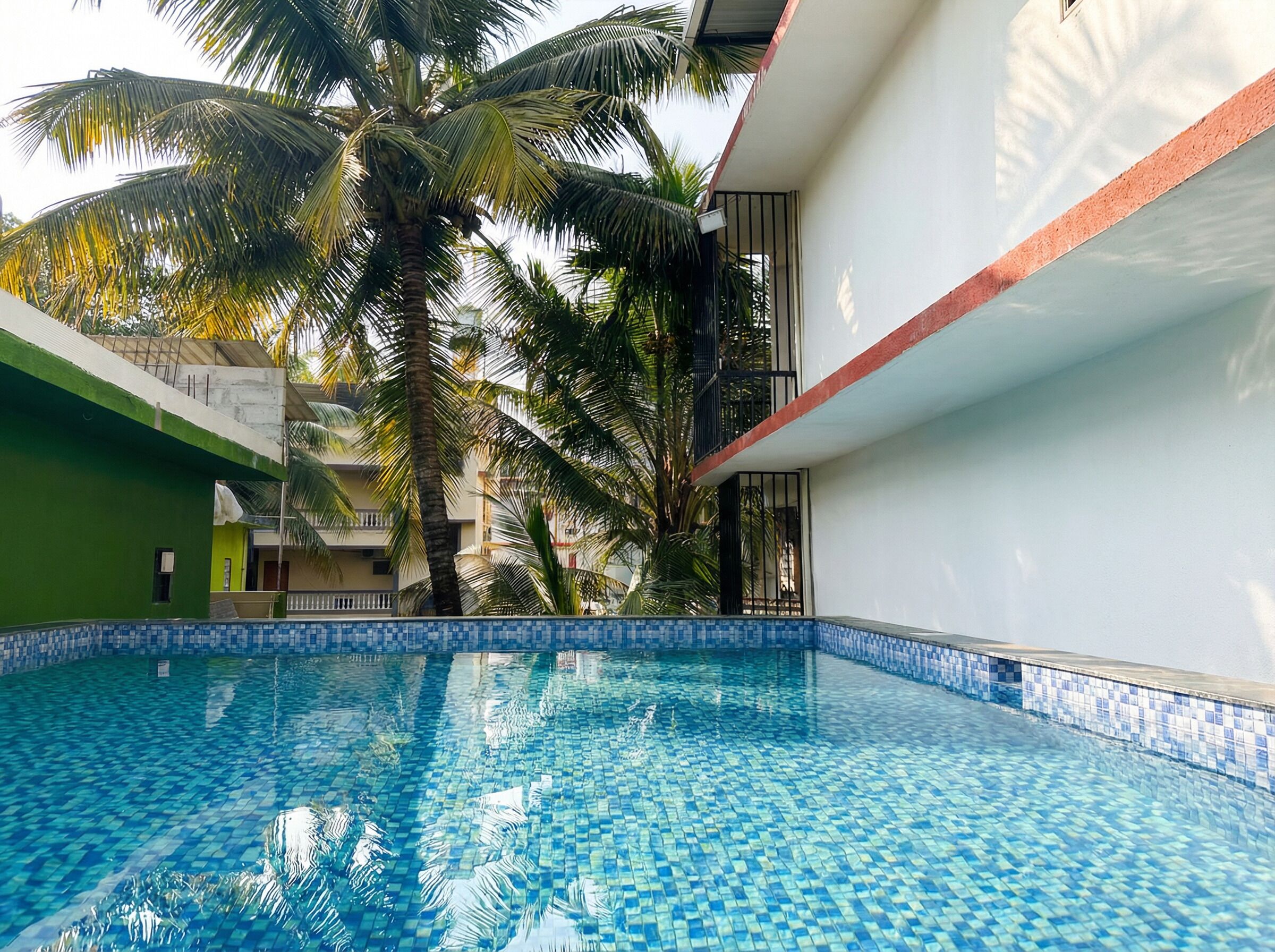 A swimming pool with blue mosaic tiles, surrounded by palm trees and a building with a balcony.